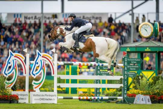 A rider jumps over a colourful obstacle in the CHIO Aachen stadium on a piebald horse. The obstacle is decorated with the horse's head from the CHIO Aachen logo on one side and represents the other Rolex Grand Slam venues, The Dutch Masters, Spruce Meadows and CHI de Genève, on the other side, which are indicated on green signposts next to the jump. In the background, numerous spectators are seated in the stands.