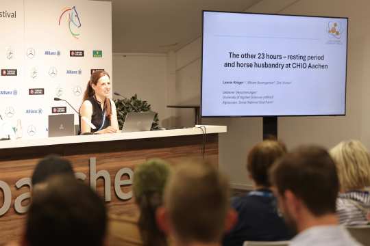 A woman sits at a lectern with a laptop and microphone and gives a presentation to the audience. The presentation entitled “The other 23 hours - resting period and horse husbandry at CHIO Aachen” can be seen on a large screen. Sponsor logos and the CHIO Aachen logo are visible in the background.