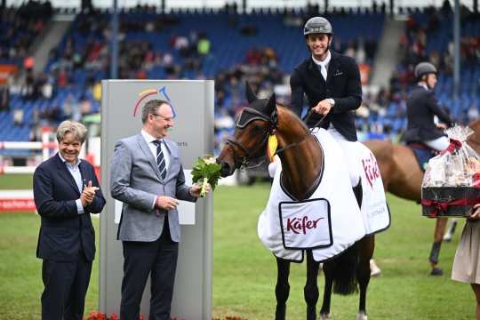 Sieger Richard Vogel in der Siegerehrung wo sein Pferd eine weiße Siegerdecke trägt. Das Pferd riecht an dem Blumenstrauß und zwei offzielle stehen daneben