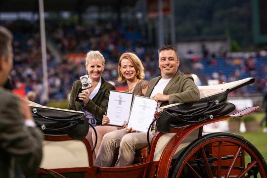 Das Foto zeigt zwei Frauen und einen Mann in einer Kutsche, die stolz ihre Preise zeigen und dabei in die Kamera lächeln.