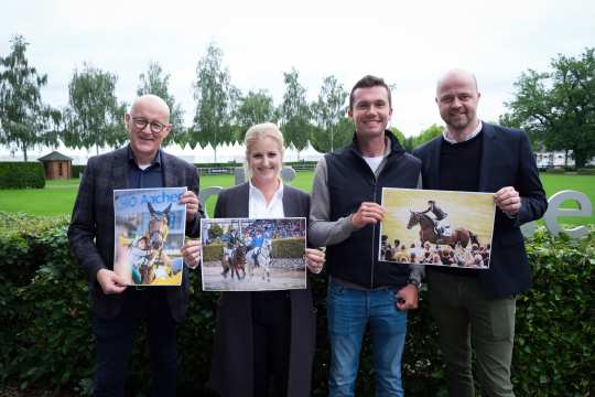 Vier Personen stehen nebeneinander im Freien und halten jeweils Fotos in den Händen. Die Personen stehen vor einer Hecke, im Hintergrund sind Bäume und ein Gebäude zu sehen. Von links nach rechts hält die erste Person ein Foto eines Pferdes mit dem Schriftzug "CHIO Aachen", die zweite Person ein Foto eines Pferdes im Sprung, die dritte Person ein Foto eines Pferdes in einer Menschenmenge und die vierte Person ein Foto eines reitenden Reiters im Gelände. Alle vier Personen lächeln in die Kamera.