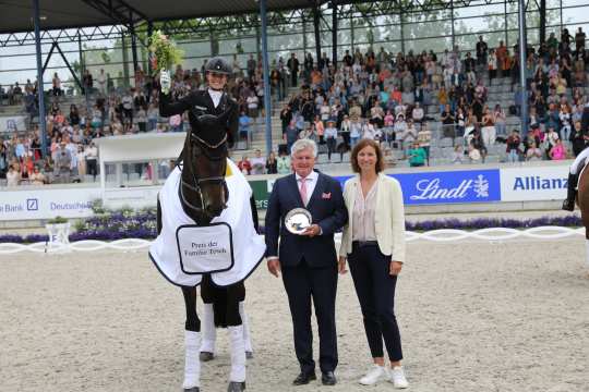 Das Foto zeigt die Dressurreitern Jessica von Bredow-Werndl auf ihrer dunkelbraunen Stute Dalera. Die Stute trägt eine weiße Abschwitzdecke und weiße Bandagen. An ihrem Kopf trägt Dalera eine goldene Schleife. Die Reiterin lächelt freundlich in die Kamera. Neben ihr stehen zwei weitere Personen, die ebenfalls in die Kamera. Im Hintergrund ist eine volle Zuschauertribüne zuerkennen.