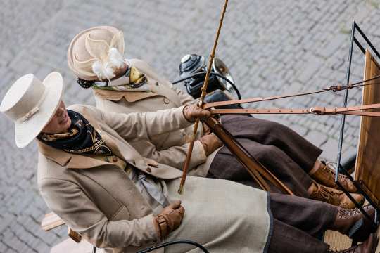 Two women in traditional dress photographed diagonally from above on the coach box of an old traditional carriage. Both are wearing a beige hat, a blue and gold scarf, a beige blazer with dark brown linen trousers and brown and cognac-coloured boots.