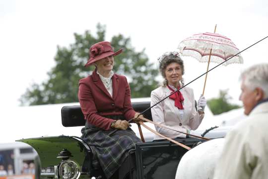 Two women in traditional dress on the coachman's seat of an old black traditional carriage. The woman on the left is wearing a bordeaux red blazer, a matching hat and has a dark chequered blanket on her legs. The woman on the right is wearing a white blazer, an old rose-coloured bow around her neck, while she holds a pink and white lace umbrella in her left hand with white lace gloves.