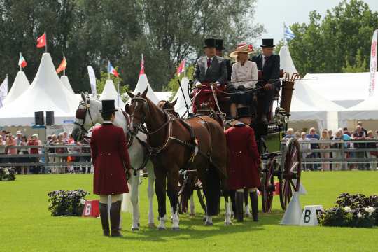 Traditioneller Vierspänner in der Mitte des Fahrstadions mit drei braunen Pferden sowie einem Schimmel vorne links. Vor und neben den Pferden stehen je ein Mann in rotem Frack, beigen Hosen und braunen Stiefeln sowie einem dunklen Zylinder. Auf dem Kutschbock sitzen neben dem Fahrer noch zwei weitere Männer und Frauen, alle traditionell gekleidet mit Zylinder oder Hut.