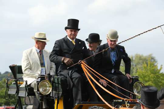 Vier Herren auf dem Kutschbock einer traditionellen Kutsche. Der Kutscher, vorne links, hält die Zügel und die Peitsche des Vierspänners in seiner linken Hand. Er trägt einen schwarzen Zylinder, Frack und Anzughose. Der Herr rechts trägt einen dunklen Nadelstreifenanzug mit schwarz-gelber Fliege und einen beigen Hut. Die Herren dahinter tragen einen beigen Anzug und Hut auf der linken Seite und einen schwarzen Anzug und Hut auf der rechten Seite.