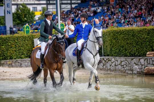 Ein Springreiter links auf einen braunen Pferd und ein anderer rechts auf einem Schimmel traben glücklich Hand in Hand durch das Wasser im Hauptstadion.