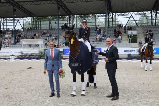 Die Reiterin Charlotte Dujardin auf ihrer dunkelbraunen Stute Times Kismet steht im Deutsche Bank Stadion und lächelt in die Kamera Neben ihr stehen zwei Männer, die ebenfalls freundlich in Richtung Kamera schauen. Das Pferd trägt eine dunkelblaue Abschwitzdecke und eine goldene Schleife am Kopf. Im Hintergrund sind viele Zuschauer auf einer Tribüne.