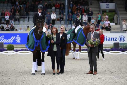Die Dressurreiterin Jessica von Bredow-Werndl sitzt auf ihrer dunkelbraunen Stute Dalera im Dressurstadion und lächelt in die Kamera. Dalera trägt eine dunkelgrüne Abschwitzdecke und weiße Bandagen. Neben ihr stehen drei weitere Personen, die ebenfalls in die Kamera lächeln. Im Hintergrund ist eine volle Zuschauertribüne zuerkennen.