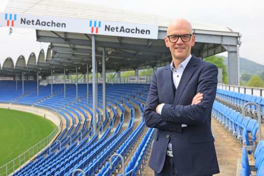 Das bild zeigt einen Herrn im blauen Anzug vor blauen Stuhlreihen in einem Reitstadion. Im Hintergrund ist das NetAachen Logo zu sehen.