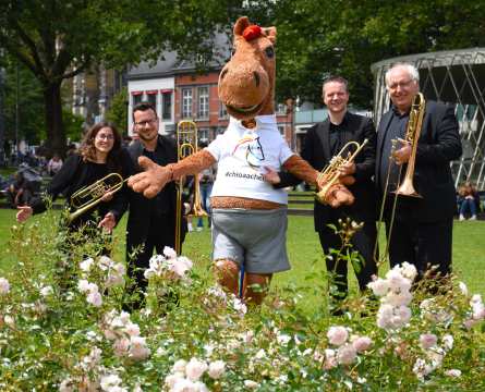 Das Foto zeigt ein etwa 2 Meter großes Maskottchen. Es ist ein Pferd, welches ein Tshirt und eine kurze Hose trägt und eingerahmt ist von schwarz gekleideten Menschen mit Musikinstrumenten.