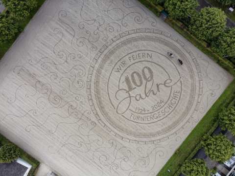 Das Bild zeigt einen kunstvoll verzierten Sandplatz aus der Vogelperspektive. Es wurde ein Logo in den sand gezeichnet mit der Inschrift "100 Jahre Turniergeschichte"