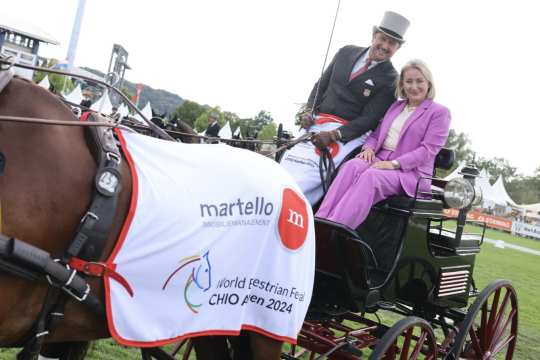 Ein Pferde zieht eine elegante Kutsche, auf der Chester Weber, der Sieger im Preis der Martello Immobilienmanagement GmbH & Co KG, sitzt. Neben ihm sitzt in einem lilafarbenen Anzug Charline Hammer, Tochter von Wolfgang (Tim) Hammer, Geschäftsführer der Martello Immobilienmanagement GmbH & Co. KG. Das Pferd trägt eine weiße Siegerdecke mit der Aufschrift „World Equestrian Festival CHIO Aachen 2024“ sowie dem Logo des Sponsors. Im Hintergrund sind Zelte, Publikum und der Rasenplatz zu sehen.
