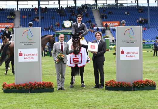 Nicola Philippaerts sitzt auf seinem braunen Pferd, welches die Siegerdecke trägt, auf der das Logo der Sparkasse Aachen abgebildet ist. Links neben dem Pferd steht ALRV-Aufsichtsratsmitglied Thomas Förl mit einem Blumenstrauß. Auf der rechten Seite des Pferdes steht der Vorstandsvorsitzende der Sparkasse Aachen Norbert Laufs, der eine Sparkassen-Tüte in der Hand hält. Im Hintergrund ist das Hauptstadion mit Tribünen und Zuschauern zu sehen.