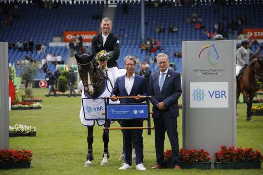 Koen Vereecke mit seinem Helm unterm Arm und einem Blumenstrauß in der Hand auf seiner braunen Stute Oilily de Muze mit ihrer weißen Siegerdecke bei der Siegerehrung in der Mitte des Hauptstadions. Rechts daneben Ralf Hündgen, Partner der Kanzlei VBR in einem blauen Anzug mit einem blauen CHIO Aachen und VBR Schriftzug zwischen braunen Holzstangen in der Hand. Daneben ALRV-Vizepräsident Frank Kemperman, ebenfalls im blauen Anzug.