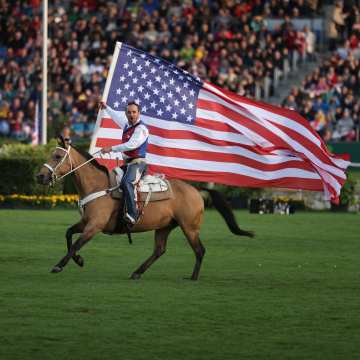 Ein im Cowboy galoppiert auf seinem Westernpferd durch Hauptstadion und trägt dabei eine riesige Amerika Flagge.