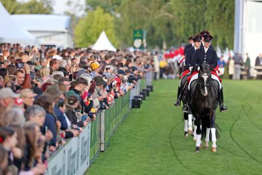 Links sind Mengen an Zuschauern und rechts reiten mehrere dunkele Pferde hintereinander ins Stadion rein.