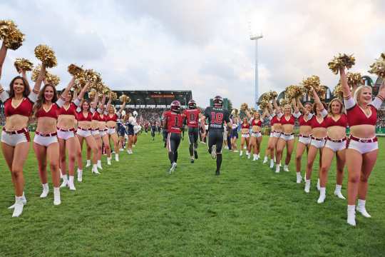 Drei Footballer rennen durch eine Gasse von Cheerleadern in roten und weißen Outfits im Hauptstadion bei der Eröffnungsfeier.