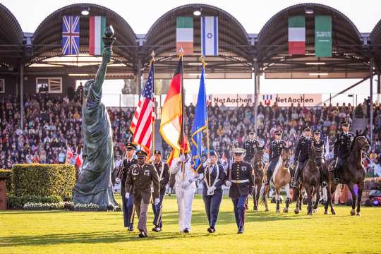 Soldiers carry the flags of the USA and Germany through the Main Stadium, accompanied by other soldiers on horseback and an imitation of the Statue of Liberty in the background.