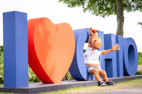 CHIO Aachen Maskottchen Karli sitzt winkend im C des großen I love CHIO-Selfiespots mit blauem I und blauen CHIO Buchstaben sowie einem roten Herz dazwischen