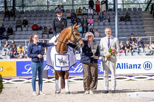 Lana-Pinou Baumgürtel auf ZINQ Emma FH NRW in der Siegerehrung der Liselott und Klaus Rheinberger Stiftung im Dressurstadion. Das Pferd trägt eine weiße Siegerdecke und zwei offizielle übergeben einen Blumenstrauß und ein Gefäß.