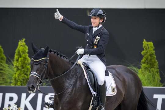Isabell Werth reitet im Schritt aus dem Stadion heraus auf ihrem schwarzen Pferd Wendy de Fontaine und zeigt ein Daumen hoch ins Publikum.