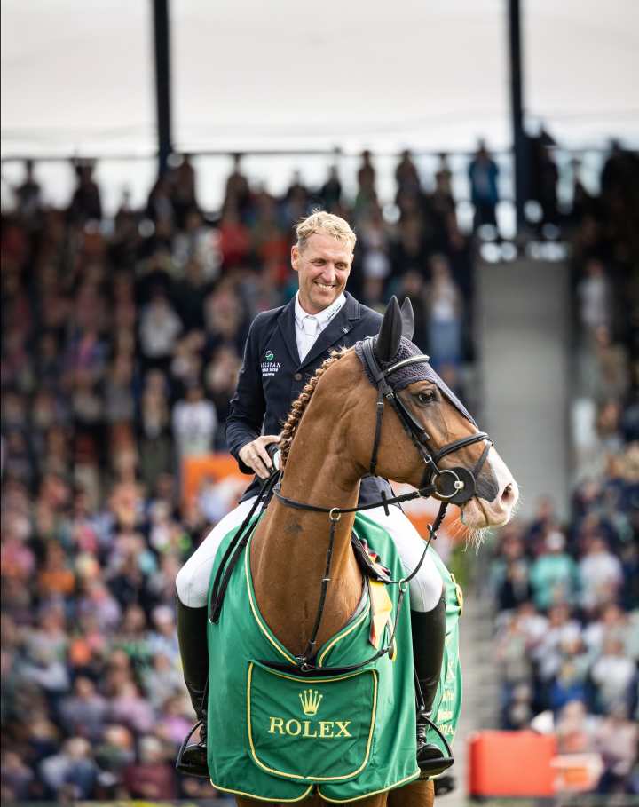 Andre Thieme lächelt auf seine Fuchsstute Chakaria herab, die ihre grüne Rolex-Siegerdecke trägt und zur Seite schaut, während er bei der Siegerehrung des Rolex Grand Prix 2024 im ausverkauften Hauptstadion im Sattel sitzt und seinen Helm abgenommenen Helm vor sich hält