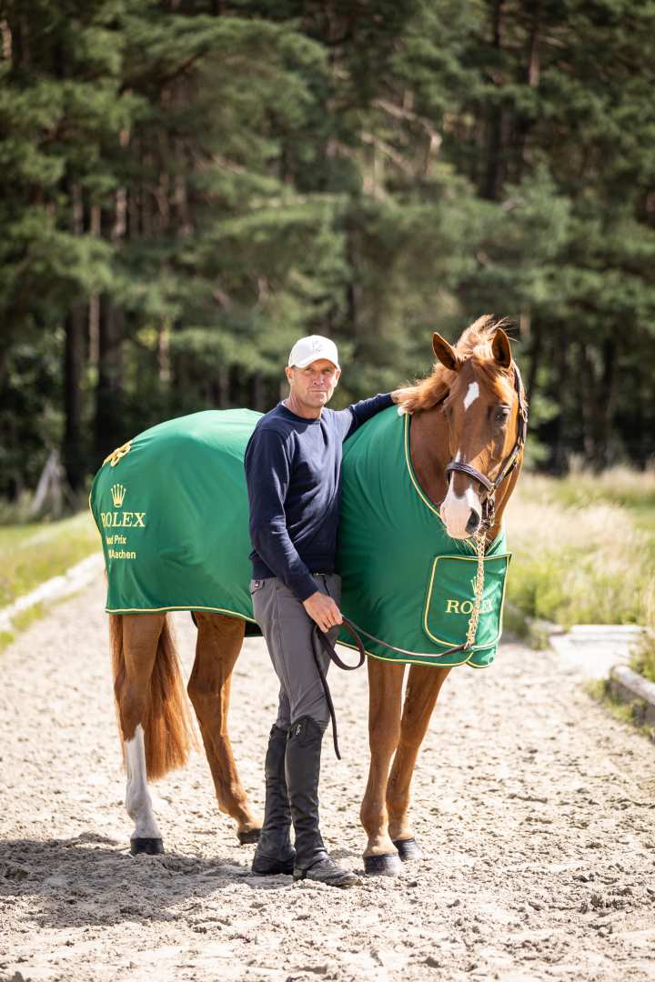 Andre Thieme mit einer weißen Kappe mit dem Emblem des Rolex Grand Slam of Show Jumping und einem Arm auf dem Rücken seiner Fuchsstute Chakaria, die ihre Rolex Grand Prix Siegerdecke trägt