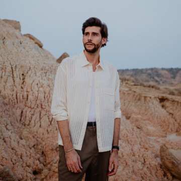 Alvaro Soler, dressed in an open, cream-coloured striped shirt over a white T-shirt and brown trousers, stands between rough rock formations against a pale sky and gazes thoughtfully into the distance.