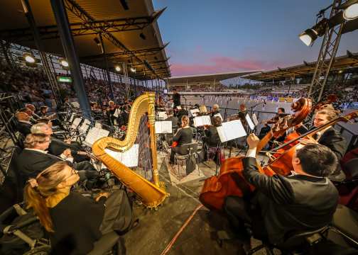 The Aachen Music Orchestra at the Horse and Symphony concert in the dressage stadium at dusk. There are many performers in the stadium and the stands in the audience are full.