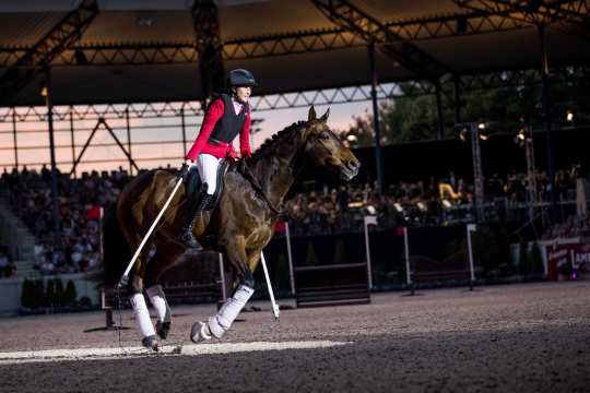 Lisa Röckener und ihr braunes Pferd Valoo in der Mitte des Dressurstadions bei ihrem Aufritt bei „Pferd & Sinfonie“. Lisa reitet nur mit Reitpad und ohne Trense, während sie zwei lange weiße Stäbe in der Hand hält, zwischen deren Enden eine Lichterkette hängt, die sie wie ein Springseil verwendet, während Valoo darüber galoppiert.