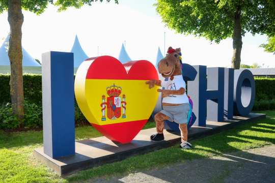 The CHIO Aachen mascot, Karli, a cheerful horse in a white T-shirt and grey trousers, stands in front of a large sculpted figure depicting the words ‘I Love CHIO’. The heart in the middle is designed in the colours of the Spanish flag and bears the Spanish coat of arms. Trees, bushes and white tent peaks can be seen in the background.