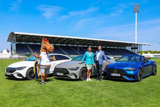 Three modern Mercedes-Benz vehicles are parked on the pitch of a stadium. A mascot in a horse costume and two elegantly dressed people pose next to the cars. A covered grandstand with the Mercedes-Benz logo can be seen in the background, with a clear blue sky above.