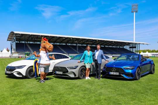 Drei moderne Mercedes-Benz Fahrzeuge stehen auf dem Rasen eines Stadions. Neben den Autos posieren ein Maskottchen in Pferdekostüm sowie zwei elegant gekleidete Personen. Im Hintergrund ist eine überdachte Tribüne mit dem Mercedes-Benz Logo zu sehen, darüber ein klarer blauer Himmel.