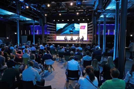 A congress in a modern remodelled industrial hall with atmospheric blue lighting. A panel discussion is taking place on the stage with four people sitting on chairs. Numerous spectators in business attire sit in the auditorium and follow the discussion attentively.