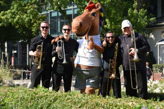 Eine Gruppe von vier Musikern in schwarzer Kleidung steht im Grünen mit Trompeten und Posaunen neben einem großen, fröhlichen Pferdemaskottchen, das ein weißes T-Shirt mit der Aufschrift #chioaachen trägt.