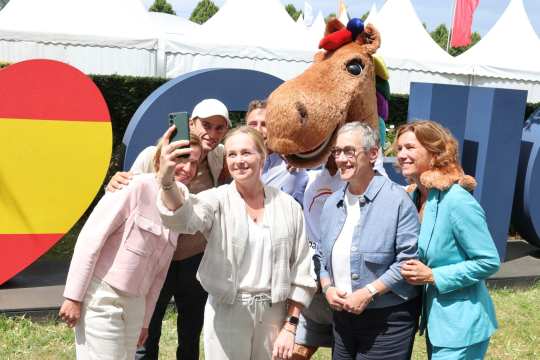 Mehrere Personen machen ein Selfie vor einer großen bunten CHIO-Buchstaben-Kulisse im Freien, begleitet von einem Maskottchen in einem Pferdekostüm mit Regenbogenmähne. Im Hintergrund sind weiße Pagodenzelte zu sehen.