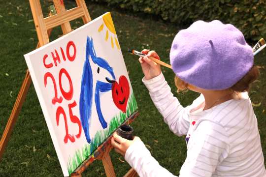 A child with a purple beret painting a colourful picture on an easel outdoors. On the canvas one can see a blue horse, a red heart with a face, a yellow sun and in red letters and figures “CHIO 2025”. The child is holding a paintbrush in the right hand and wearing a striped long-sleeved shirt.