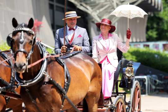 An elegantly dressed couple sits in a historic horse-drawn carriage. The man is wearing a light suit with a bow tie and a straw hat, while the woman is dressed in a pink outfit with a matching hat and holds a white parasol. Two brown horses are pulling the carriage.