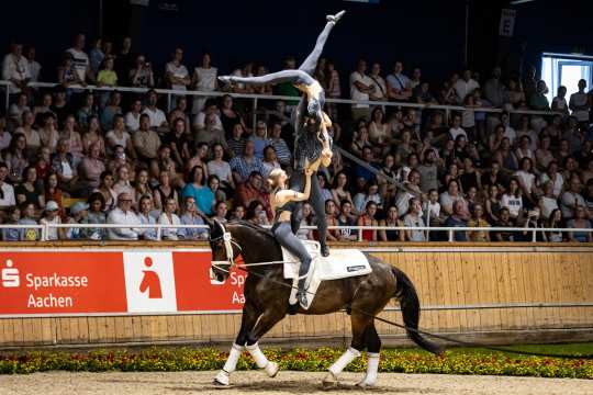 Zwei Voltigiererinnen führen eine akrobatische Kür auf einem galoppierenden Pferd in einer Arena vor. Eine Athletin steht auf dem Pferderücken und hebt die andere, die in einer eleganten Pose mit ausgestrecktem Bein in die Luft gehalten wird. Im Hintergrund sitzt das Publikum auf den Tribünen. Am Rand der Arena ist ein Schild mit der Aufschrift „Sparkasse Aachen“ zu sehen.