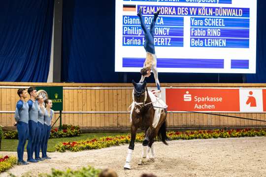 A vaulter performs a handstand on a cantering horse inside an arena, while team members in blue uniforms stand at the edge watching. In the background, a large scoreboard displays the team name and participants, alongside sponsor banners. The horse is equipped with white bandages and vaulting gear, and the scene is framed by flower beds and a wooden wall.