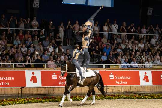 Three vaulters perform an acrobatic routine on the back of a bay horse in an indoor arena. Two people stand on the horse, while a third person strikes an elegant pose with an outstretched leg pointing upwards. Numerous spectators sit in the stands in the background. The band is decorated with red advertising banners of Sparkasse Aachen.
