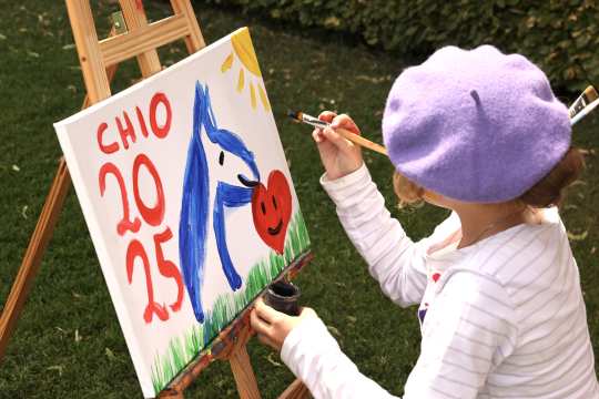 A child with a purple beret painting a colourful picture on an easel outdoors. On the canvas one can see a blue horse, a red heart with a face, a yellow sun and in red letters and figures “CHIO 2025”. The child is holding a paintbrush in the right hand and wearing a striped long-sleeved shirt.