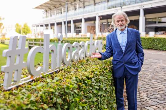 Ein Mann im blauen Anzug steht neben einer Hecke, auf der ein großes, silbernes Hashtag-Zeichen mit der Aufschrift "#chioaachen" angebracht ist. Im Hintergrund ist ein Teil des Stadions und weitere Begrünung zu sehen.