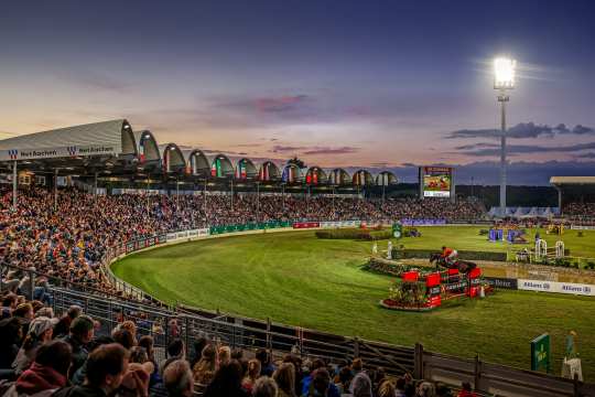 Großes Reitstadion bei Sonnenuntergang, voll besetzte Tribünen und beleuchtetes Springturnier. Im Vordergrund springt ein Reiter mit seinem Pferd über ein Hindernis, während das Publikum das Geschehen aufmerksam verfolgt.
