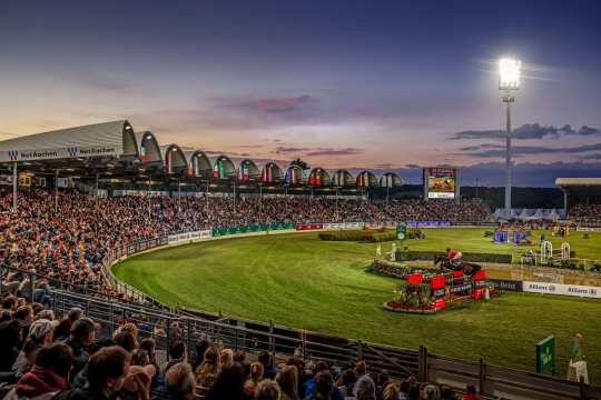 Großes Reitstadion bei Sonnenuntergang, voll besetzte Tribünen und beleuchtetes Springturnier. Im Vordergrund springt ein Reiter mit seinem Pferd über ein Hindernis, während das Publikum das Geschehen aufmerksam verfolgt.
