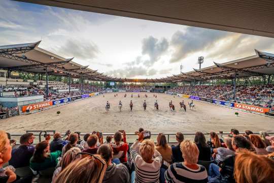 View of a large dressage stadium with full grandstands. In the centre of the arena, riders on their horses and dancers with fans can be seen forming a formation. Spectators watch the show at sunset, some taking photos of the action with their phones.