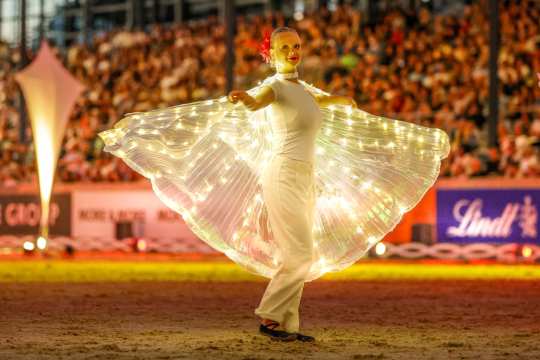 A person stands in the centre of an arena wearing a glowing costume with outstretched wings interspersed with strings of lights. The person is dressed all in white and dancing on a sandy floor, with a large, blurred crowd of people in the stands in the background.