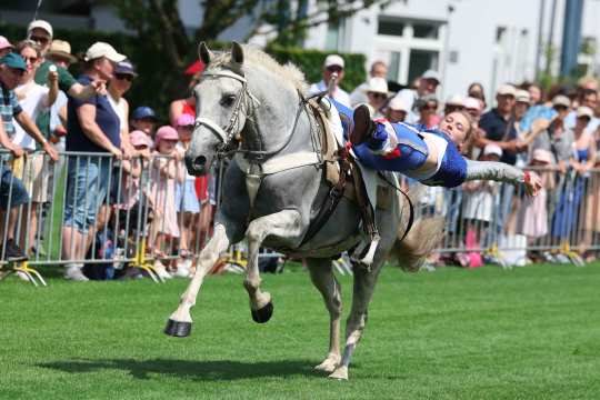 A woman in a blue glitter costume hangs horizontally from the side of a galloping grey horse at Soerser Sonntag. The audience in the background is thrilled.