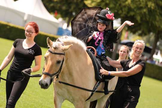 A young girl performing therapeutic riding in a costume with a black hat, lace, colourful ribbons and a black parasol sits on a Haflinger. Two adults dressed in black lead the horse and support the child from the side. White tents and trees can be seen in the background.
